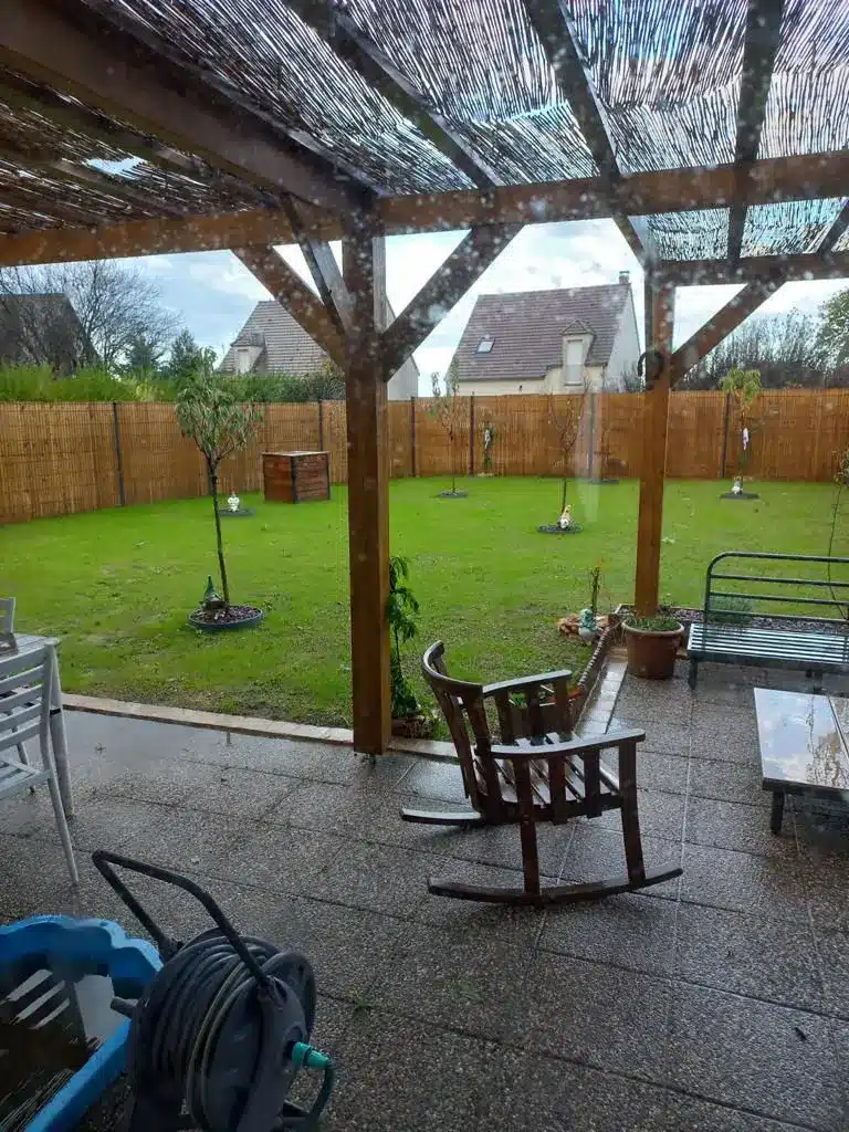 Vue sur un jardin entouré de clôture, la vue est depuis un abri de jardin. On voit qu'il pleut.